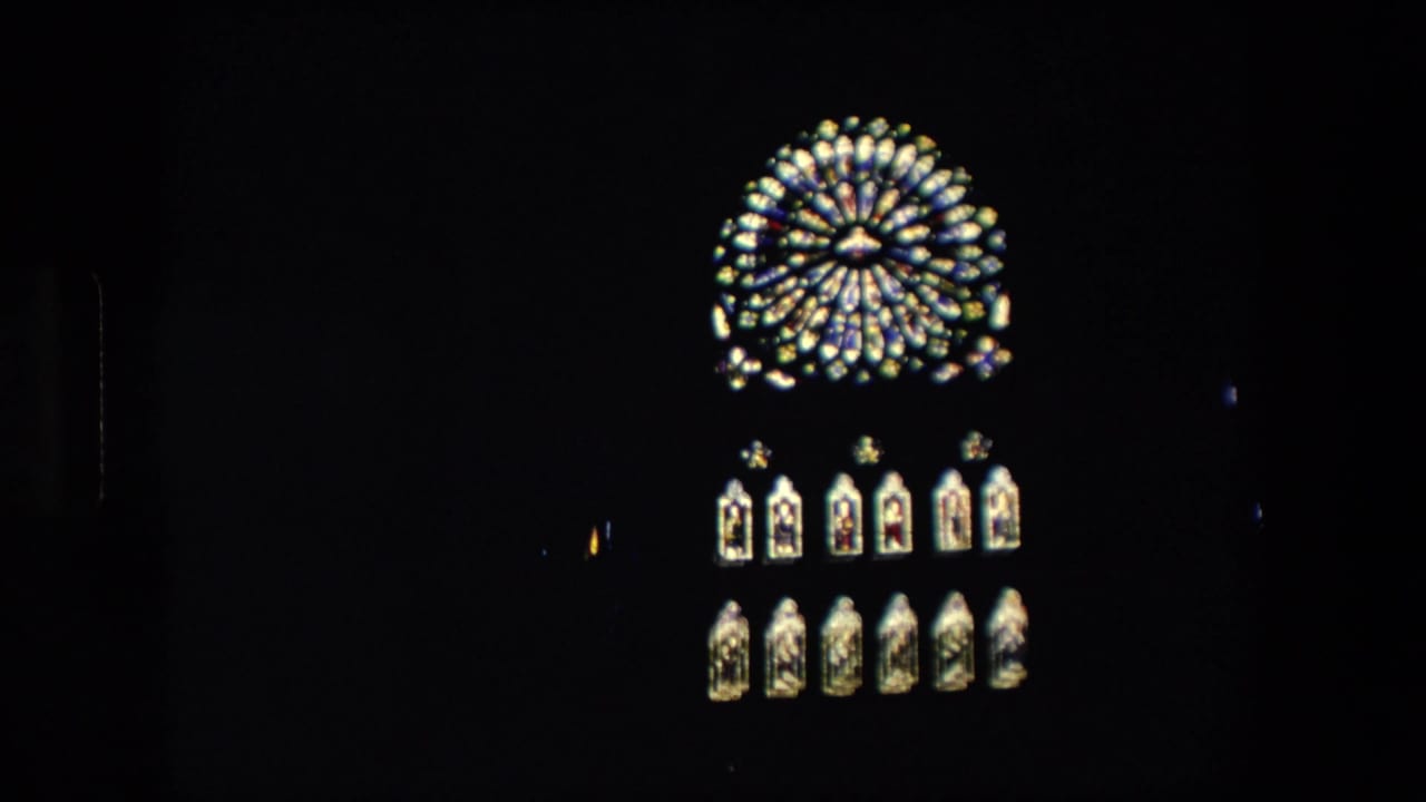 1967 London home-movie frame showing a glowing circular clock or window-like feature above lit arched panels against a nearly black nighttime facade.