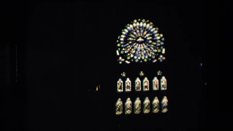 1967 London home-movie frame showing a glowing circular clock or window-like feature above lit arched panels against a nearly black nighttime facade.