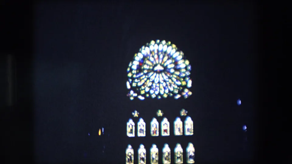 1967 London home-movie frame showing a softly focused stained-glass church window with a circular rose-like upper section and smaller panes below in a dark interior.