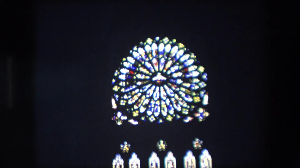 Close home-movie view of an illuminated church stained-glass window in London, with a rose-style circular pattern and small lower panels visible against deep shadow.
