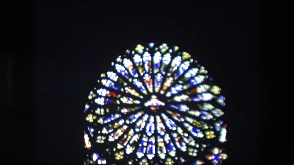 Closer 8mm frame of a bright circular stained-glass church window, with radial tracery and colored panes softened by focus and film grain.
