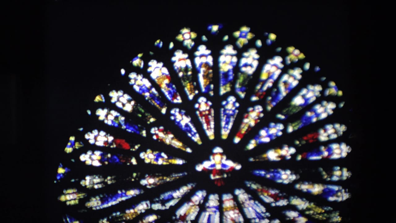 1967 London home-movie frame showing a circular stained glass window inside a Catholic church, with radiating colored panes and a small central figure.