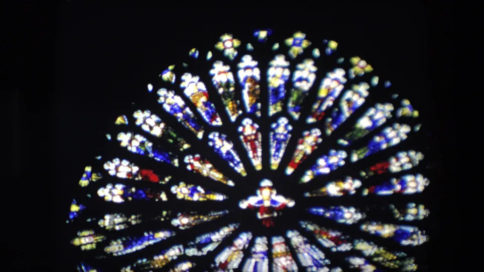1967 London home-movie frame showing a circular stained glass window inside a Catholic church, with radiating colored panes and a small central figure.