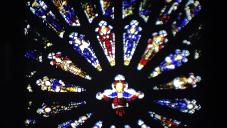 Home-movie frame of a stained glass fan or rose-window section inside the Catholic church, with saturated colored panels and a small robed figure near the bottom center.