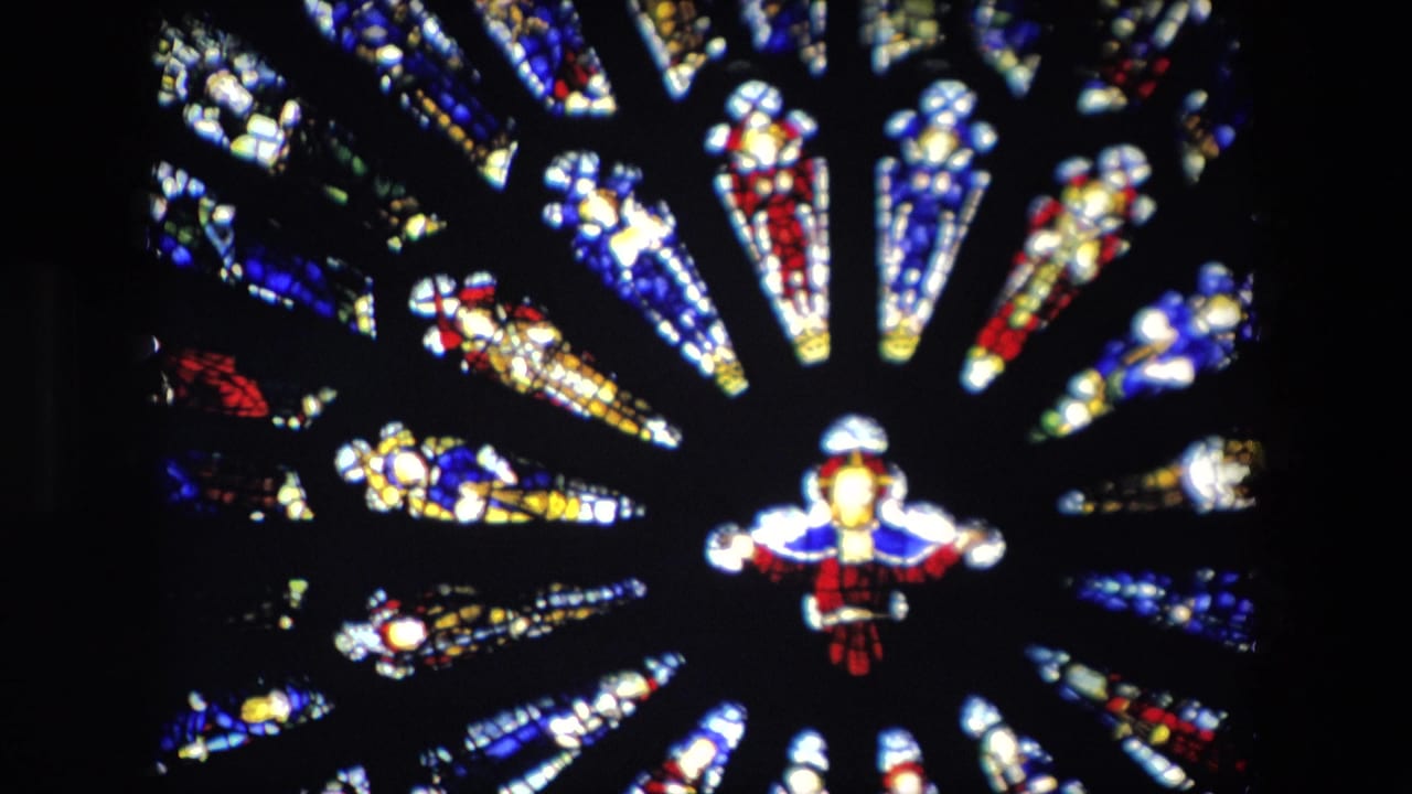 1967 London home-movie frame showing a close, slightly soft view of a circular stained glass window with red, blue, yellow, and white glass around a central figure-like motif.