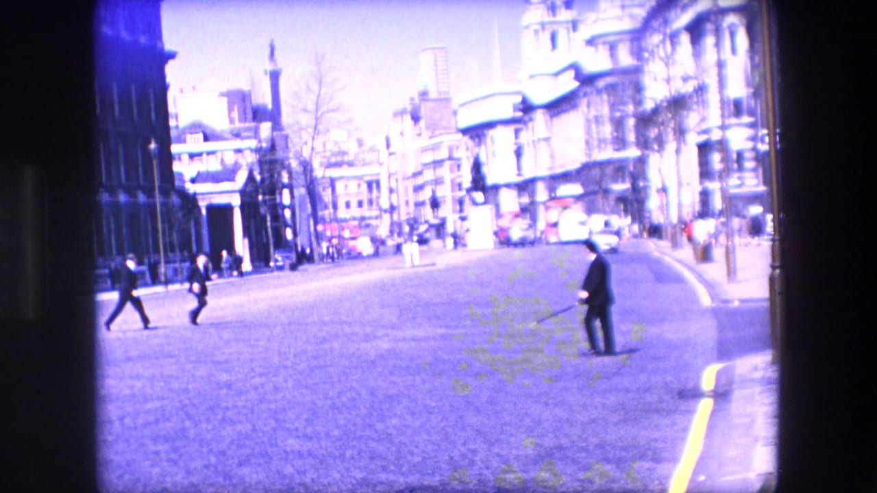Suited pedestrians cross a broad city street while cars and stone buildings line the road.