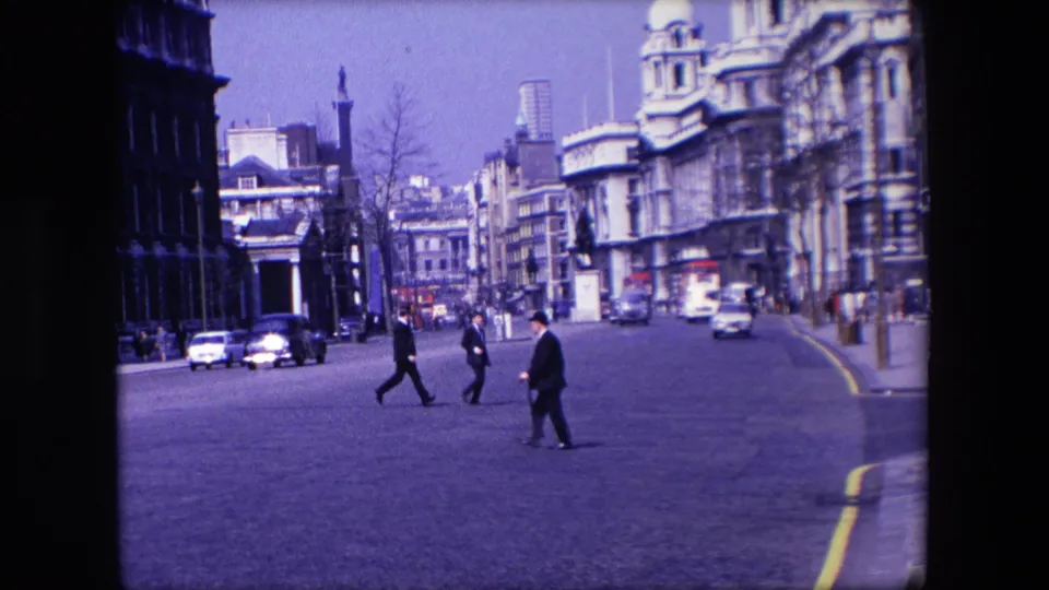 In 1967 London, men in dark suits and hats cross a broad road with moving cars, civic-looking buildings, and street furniture visible around them.