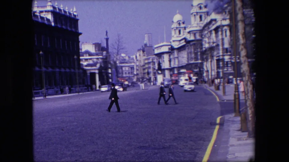 A 1967 London street view shows a suited man crossing in the foreground, with additional pedestrians, automobiles, pavements, and large stone buildings behind him.