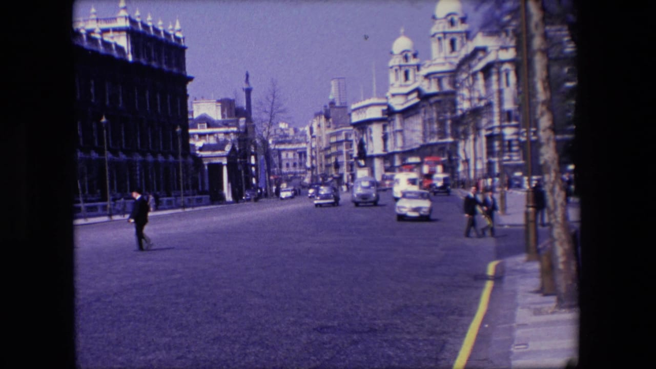 1967 London home-movie frame showing a broad city street with period cars, pedestrians crossing, tall buildings, poles, and bright daylight.