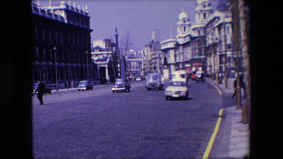 1967 London street view with cars moving toward the camera, pedestrians near the left edge, ornate building facades, and a yellow curb line.