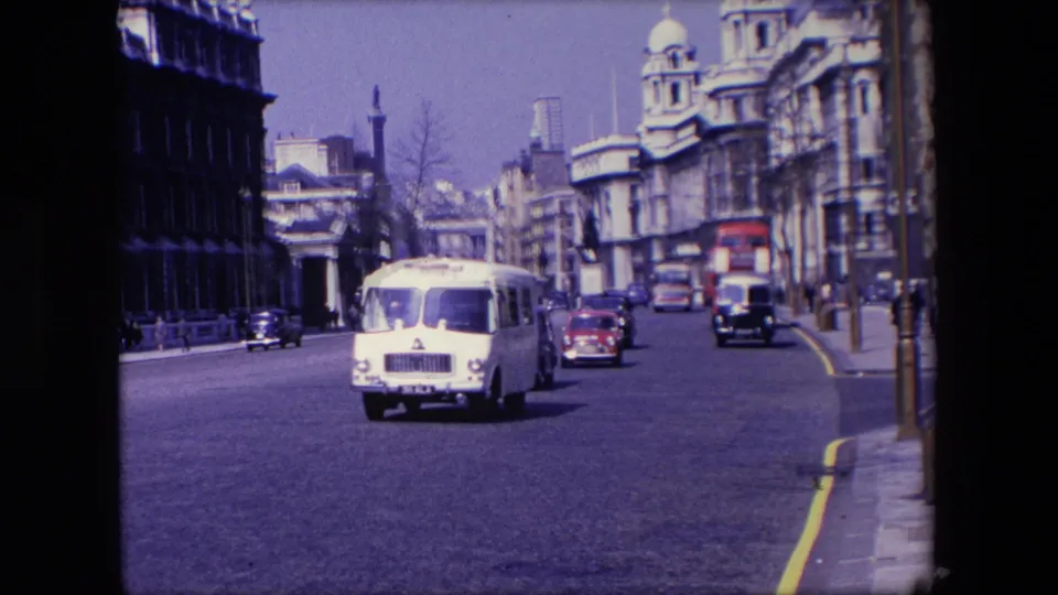 1967 London home-movie frame with a light-colored van or bus, red and dark cars, pedestrians, street poles, and prominent city building facades.