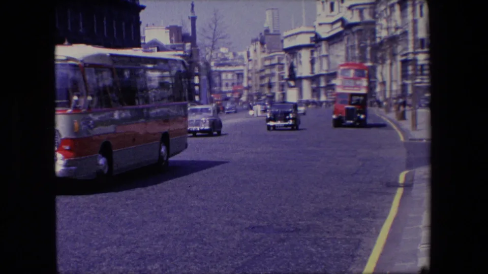1967 London traffic scene with a red and gray bus moving left of frame, cars following behind, and urban buildings along the road.