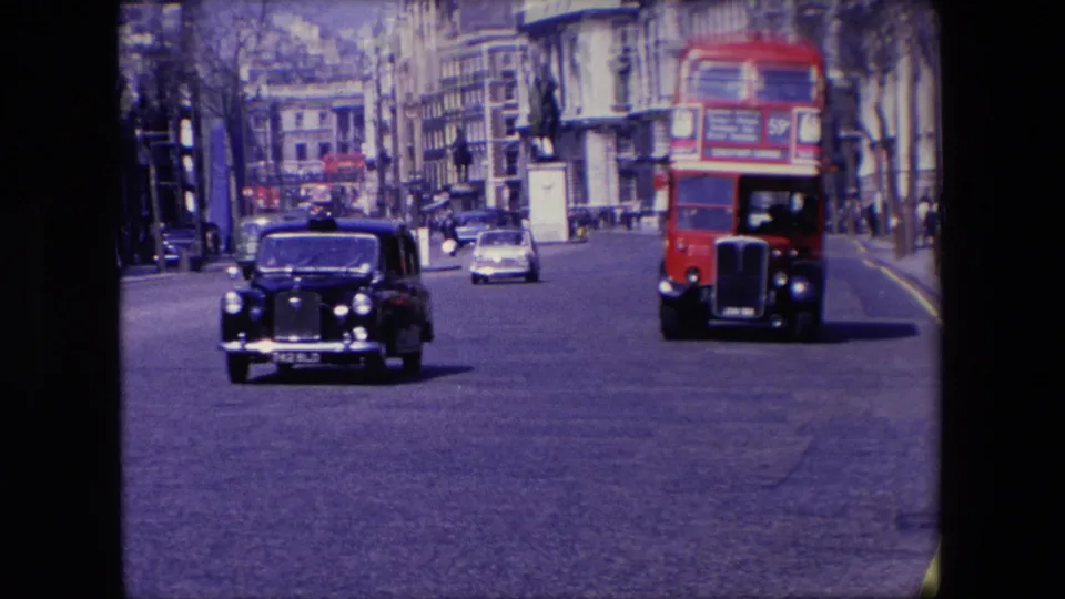 1967 London roadway with a black taxi in the foreground, a red double-decker bus approaching, and cars moving between rows of buildings.