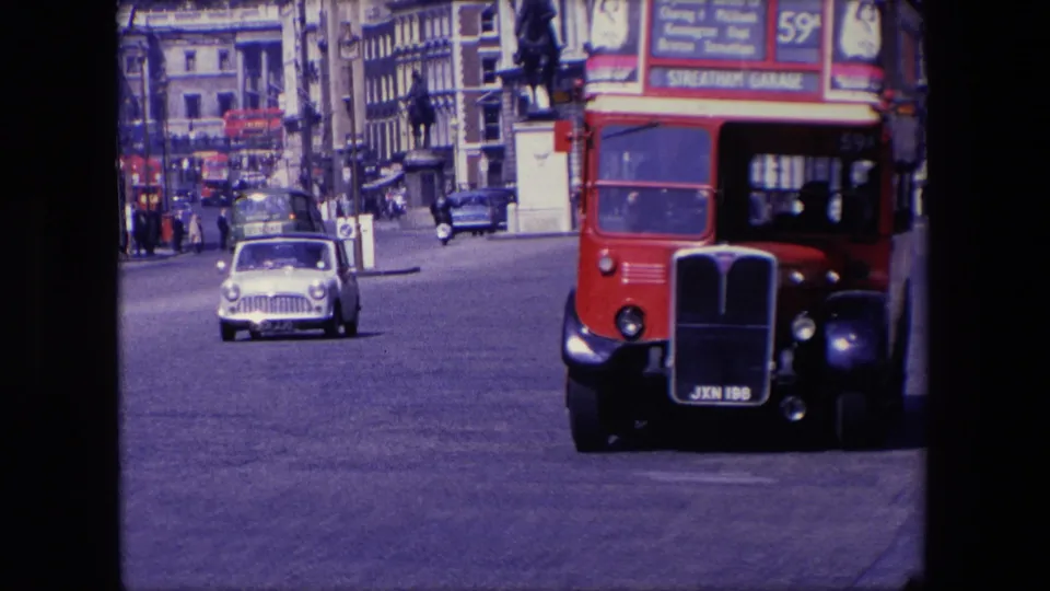 1967 London street traffic with a red double-decker bus close at right, a small white car approaching, and commercial signs on buildings in the background.