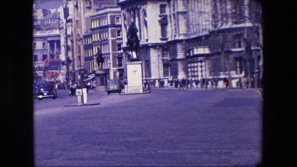 Wide 1967 London roadway view with cars in motion, pedestrians along the pavement, a traffic island, a statue or monument, and large stone-fronted buildings.
