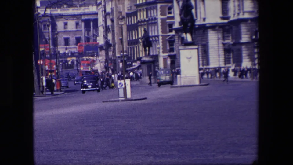 1967 London street view showing black cars, red double-decker buses in the distance, pedestrians near building fronts, and a central traffic island.