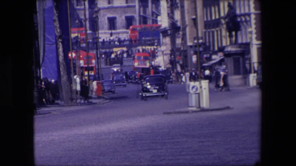 1967 London roadway frame with a black car approaching, red double-decker buses in the background, pedestrians at the street edge, and tall commercial or civic buildings around the intersection.