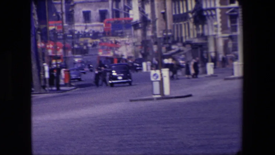 1967 London street view showing dark cars moving along a broad road, pedestrians near the pavements, red double-decker buses in the distance, and older urban buildings.