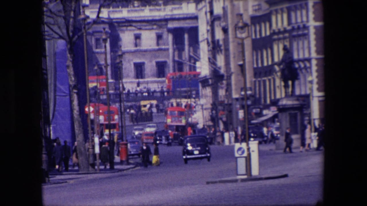 1967 London urban traffic scene with a dark saloon car approaching, red buses farther up the street, pedestrians at the sides, and mixed stone and timber-fronted buildings.