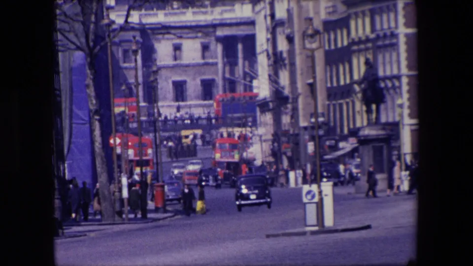 1967 London urban traffic scene with a dark saloon car approaching, red buses farther up the street, pedestrians at the sides, and mixed stone and timber-fronted buildings.