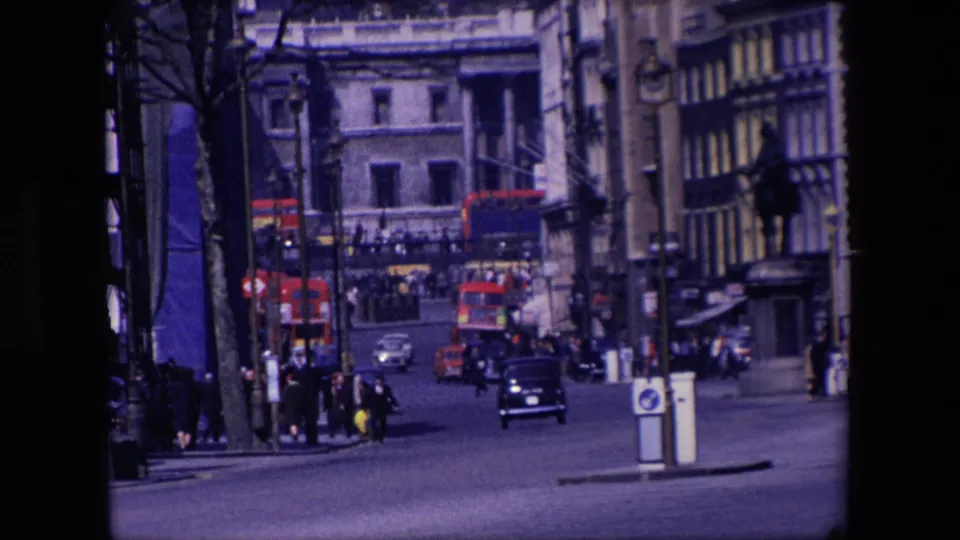 1967 London street view from a home-movie camera, with pedestrians crossing or waiting near the pavement, red buses and cars in traffic, and older buildings enclosing the roadway.