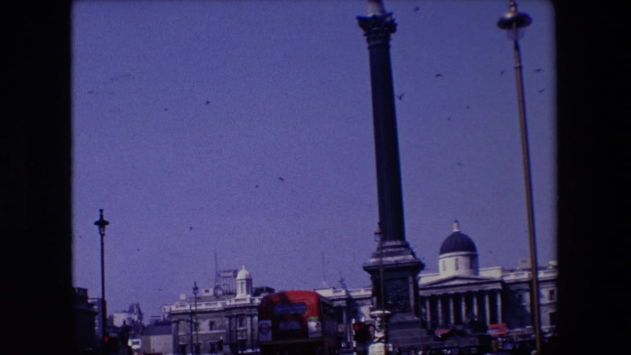 Tall column monument with birds in the sky, a domed building, street lamps, and a red bus below.