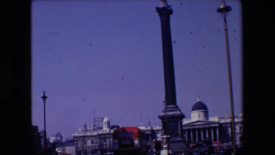 1967 London home-movie frame showing a tall statue-topped column, birds crossing a clear blue sky, classical buildings, street lamps, and a red double-decker bus near the lower edge.