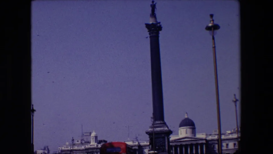 1967 London frame with the statue-topped column rising above civic buildings, a domed roof in the background, a red bus partly visible, and small birds scattered across the sky.