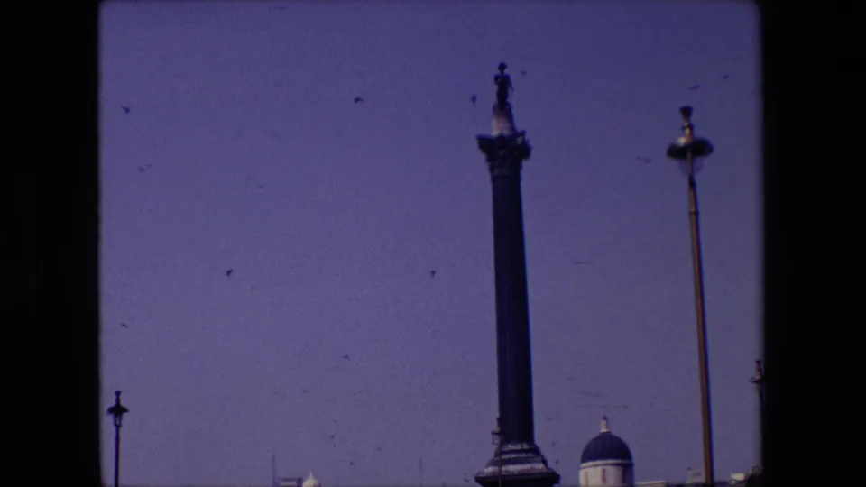 1967 London home-movie frame tilted upward toward the statue-topped column, with birds in the open sky, a tall street lamp, and the tops of classical buildings along the lower edge.