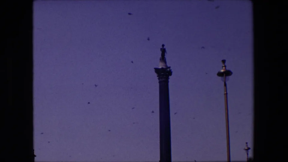 1967 London frame showing the upper portion of a tall column monument, multiple street lamps, birds scattered across a clear blue sky, and low civic buildings at the horizon.