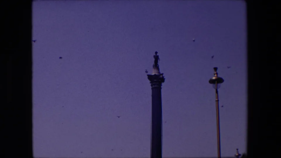1967 London home-movie frame showing a tall column-top statue, a nearby streetlamp, and small birds crossing the sky.