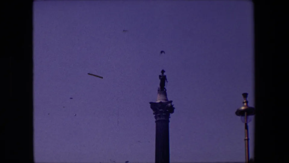A London 1967 8mm frame holds on the upper part of a tall monument as birds move across the open sky beside a streetlamp.