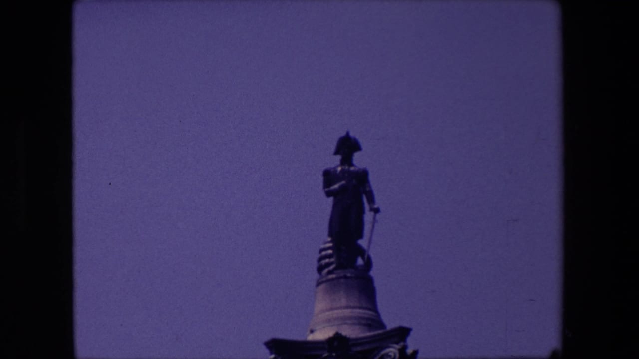 1967 London home-movie frame showing a dark standing statue on a raised pedestal against open sky, with visible 8mm frame edges.