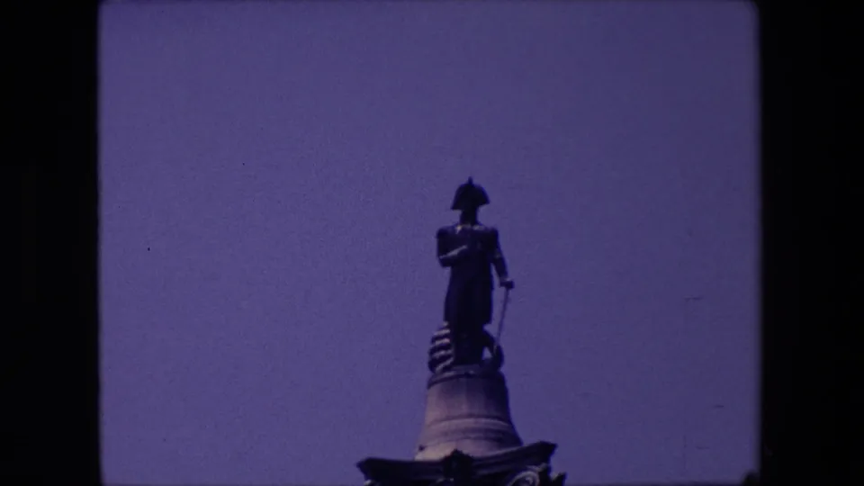 1967 London home-movie frame showing a dark standing statue on a raised pedestal against open sky, with visible 8mm frame edges.