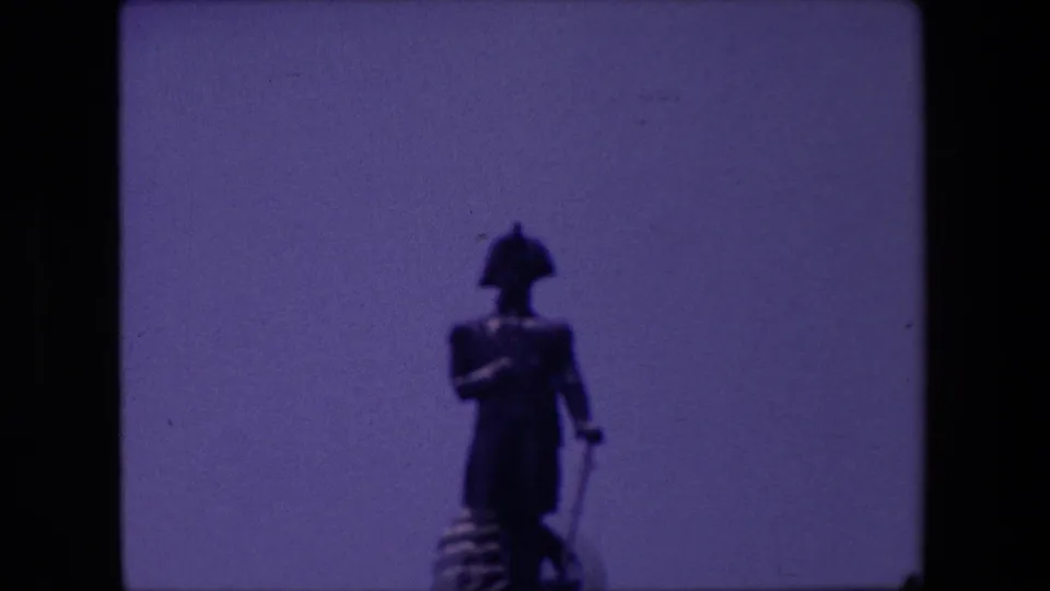 Soft 8mm frame from the supplied 1967 London clip, centered on a standing statue with a pedestal partly visible below.