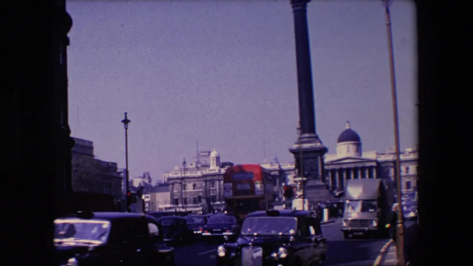 The frame shows 1967 London traffic with dark cars, a light delivery truck, tall columns, street lamps, and pale stone buildings under a bright sky.