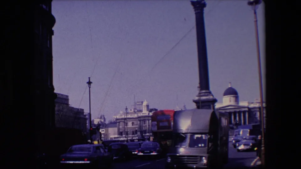A later frame in the 1967 London clip shows a delivery truck close to the camera, cars and a red bus in traffic, with stone architecture and a tall column behind them.