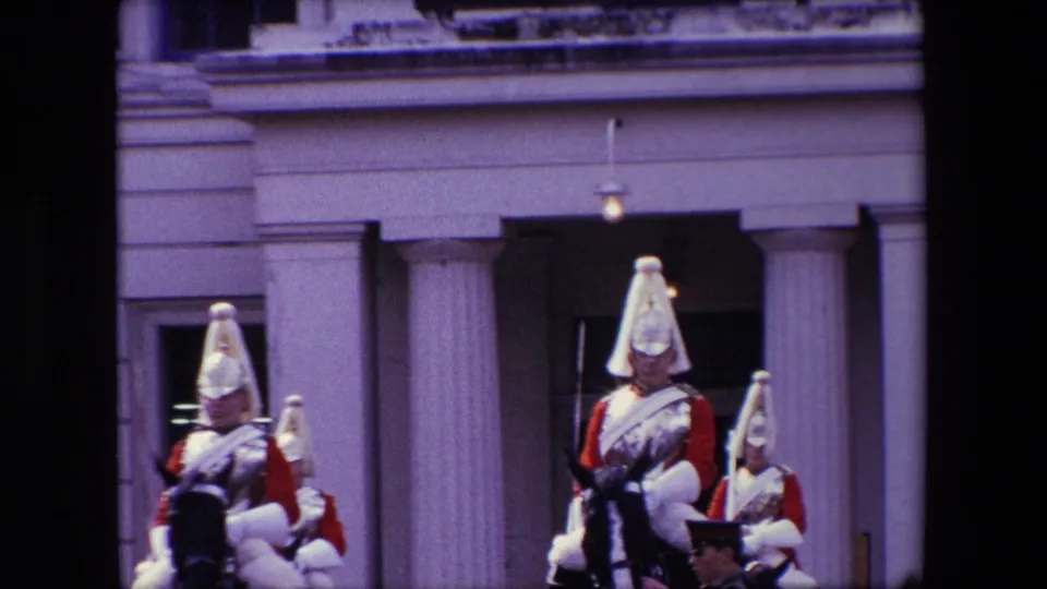 A closer 8mm frame shows mounted cavalrymen in ceremonial dress passing or presenting in front of a columned London building entrance.