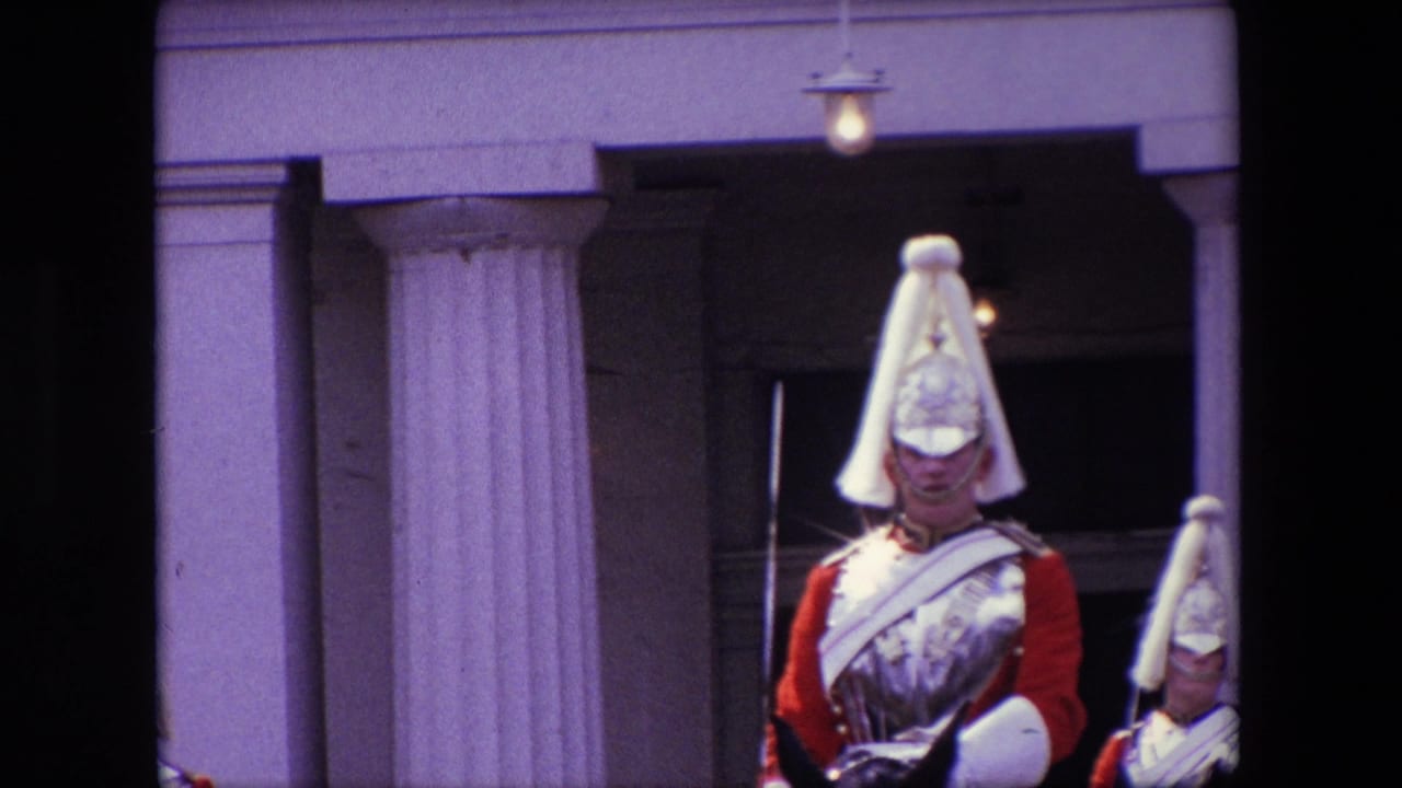 1967 London home-movie frame showing a mounted ceremonial guard in a red uniform and plumed helmet near pale stone columns, partly obscured by dark foreground edges.