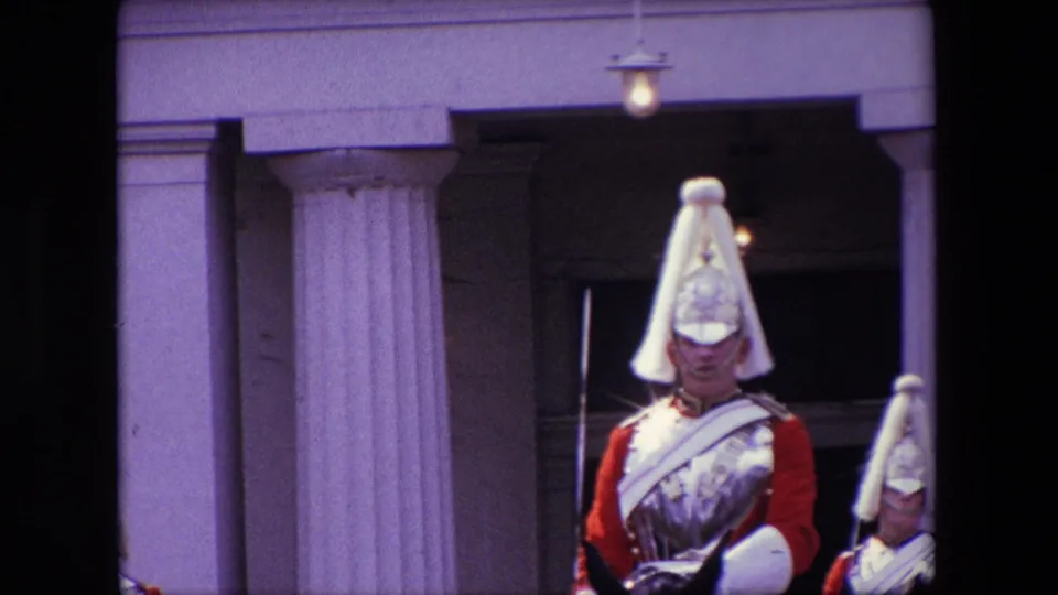 1967 London home-movie frame showing a mounted ceremonial guard in a red uniform and plumed helmet near pale stone columns, partly obscured by dark foreground edges.