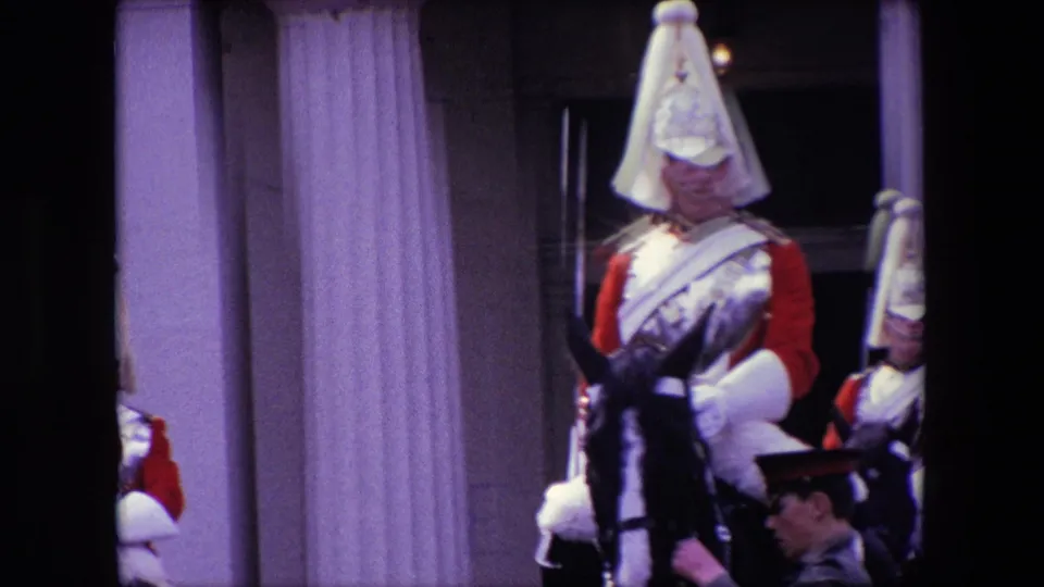 1967 London frame with mounted ceremonial guards in red uniforms and polished helmets, a black horse, and a uniformed attendant on foot in front of a columned building.