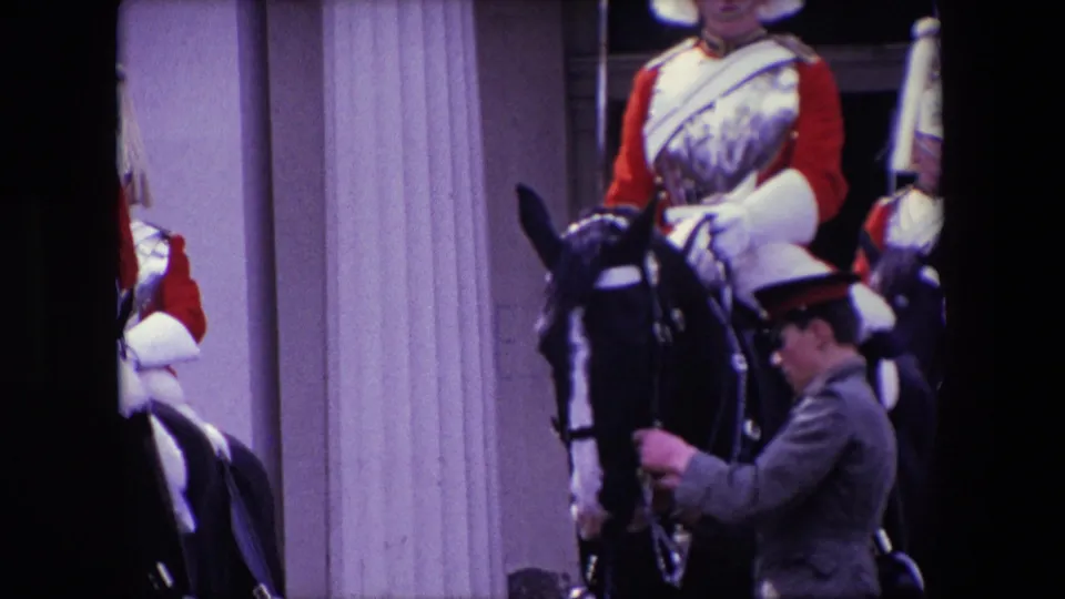 1967 London 8mm frame showing a mounted guard in red ceremonial dress and an attendant handling a black horse, with stone columns and dark foreground obstruction in view.