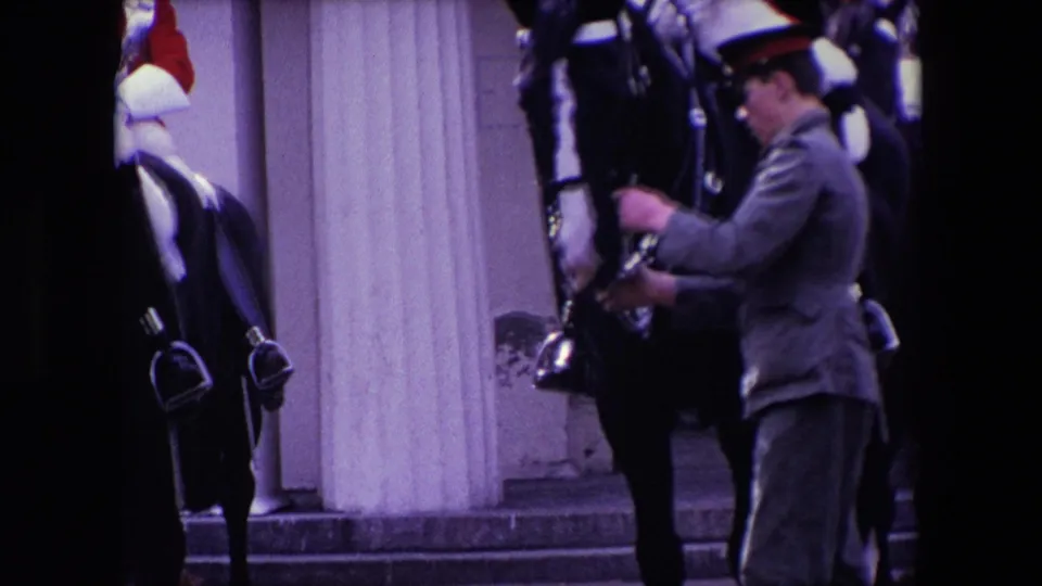 1967 London home-movie frame of a mounted ceremonial guard in red uniform and plumed helmet with a uniformed attendant standing by the horse near a formal columned facade.