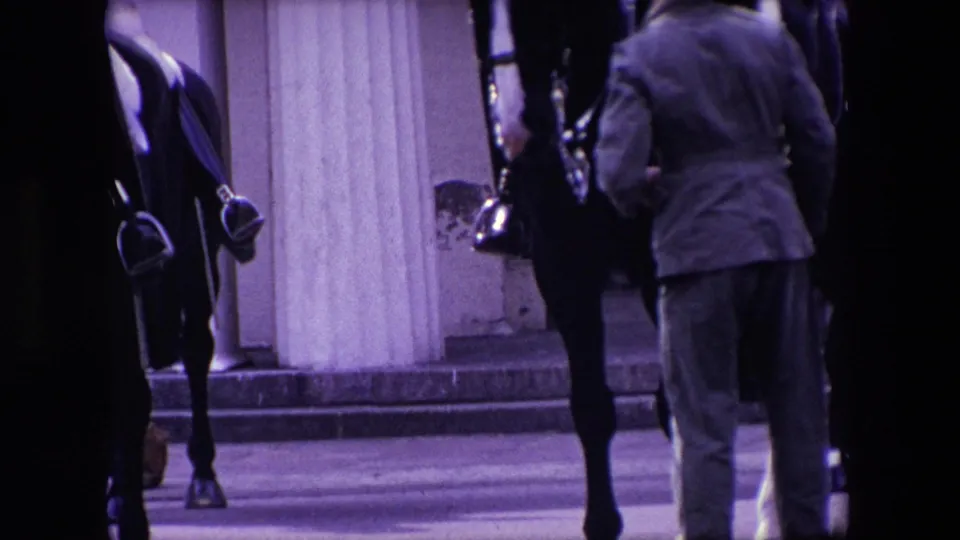 1967 London home-movie frame showing dark horses with mounted uniformed soldiers partly obscured by a man standing near stone columns.