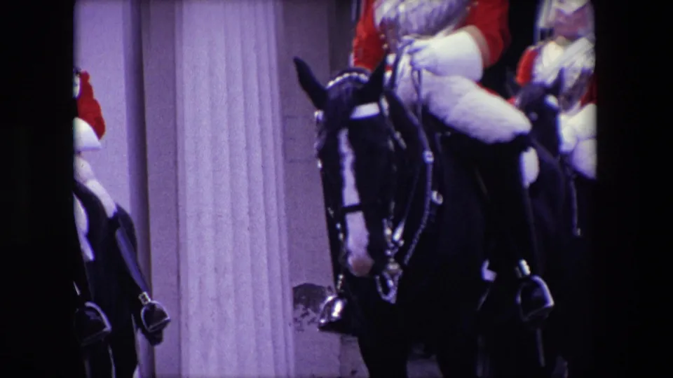 1967 London frame of a mounted ceremonial soldier with red sleeves and pale riding gear on a dark horse beside a stone column.