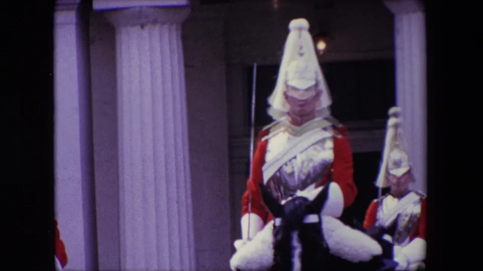 1967 London home-movie frame showing mounted uniformed soldiers wearing pale ceremonial helmets and red sleeves in front of a columned building.