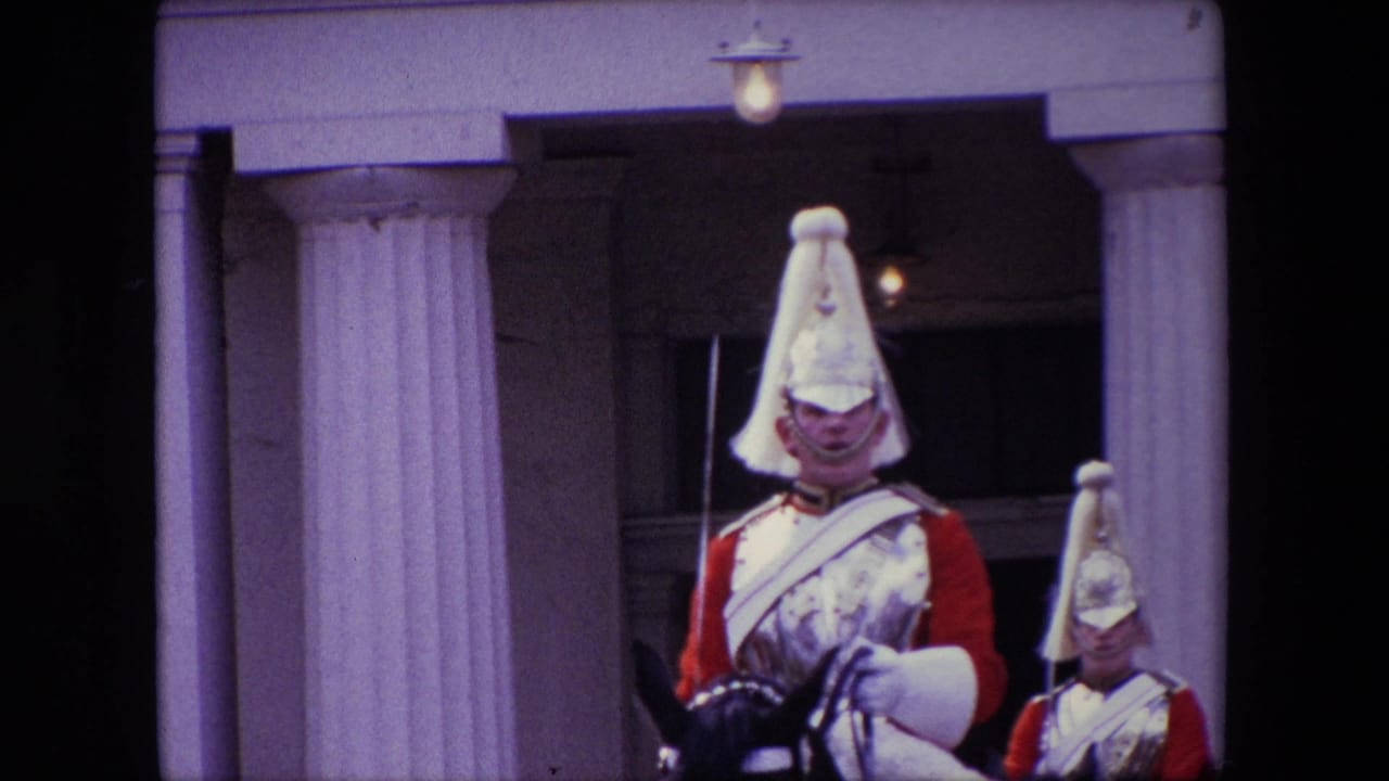 A mounted uniformed guard in a red tunic and polished helmet passes close to the camera outside a formal London building, with another mounted guard partly visible behind him.