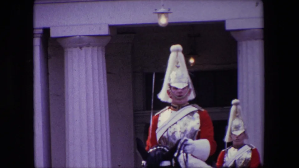 A mounted uniformed guard in a red tunic and polished helmet passes close to the camera outside a formal London building, with another mounted guard partly visible behind him.