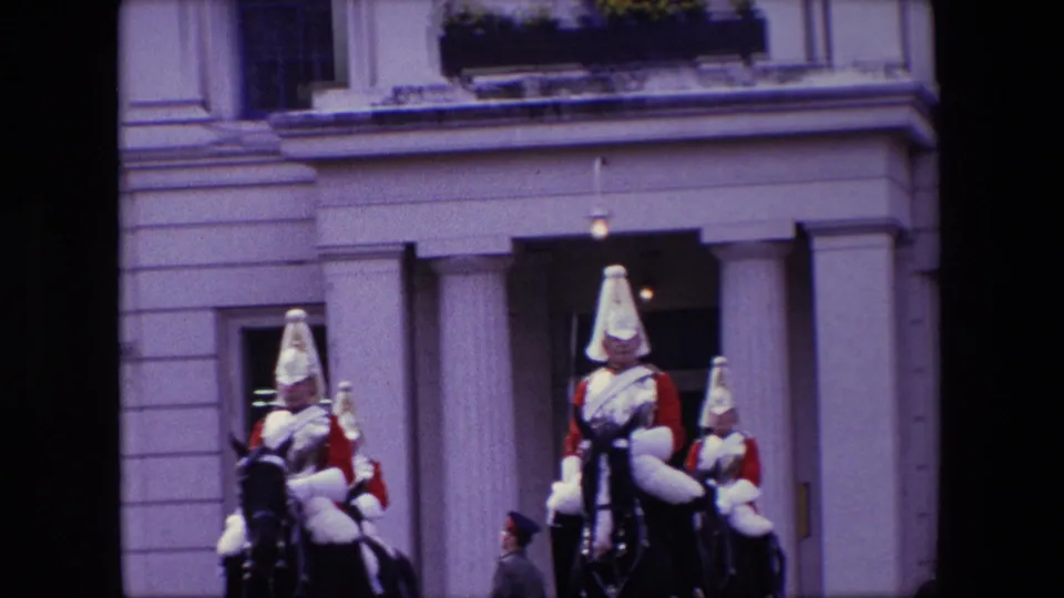 Several mounted ceremonial guards in red tunics and bright helmets ride across an open forecourt in front of a pale London building with columns and windows.