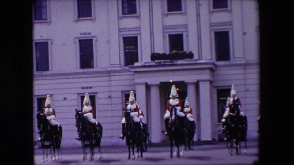 A wider 1967 London home-movie view shows mounted guards arranged across a paved forecourt before a large pale building with tall windows and a columned doorway.